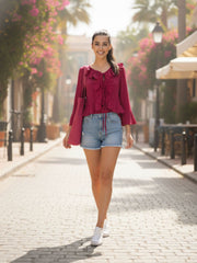 Woman walking on a sunlit street wearing a red blouse and denim shorts.