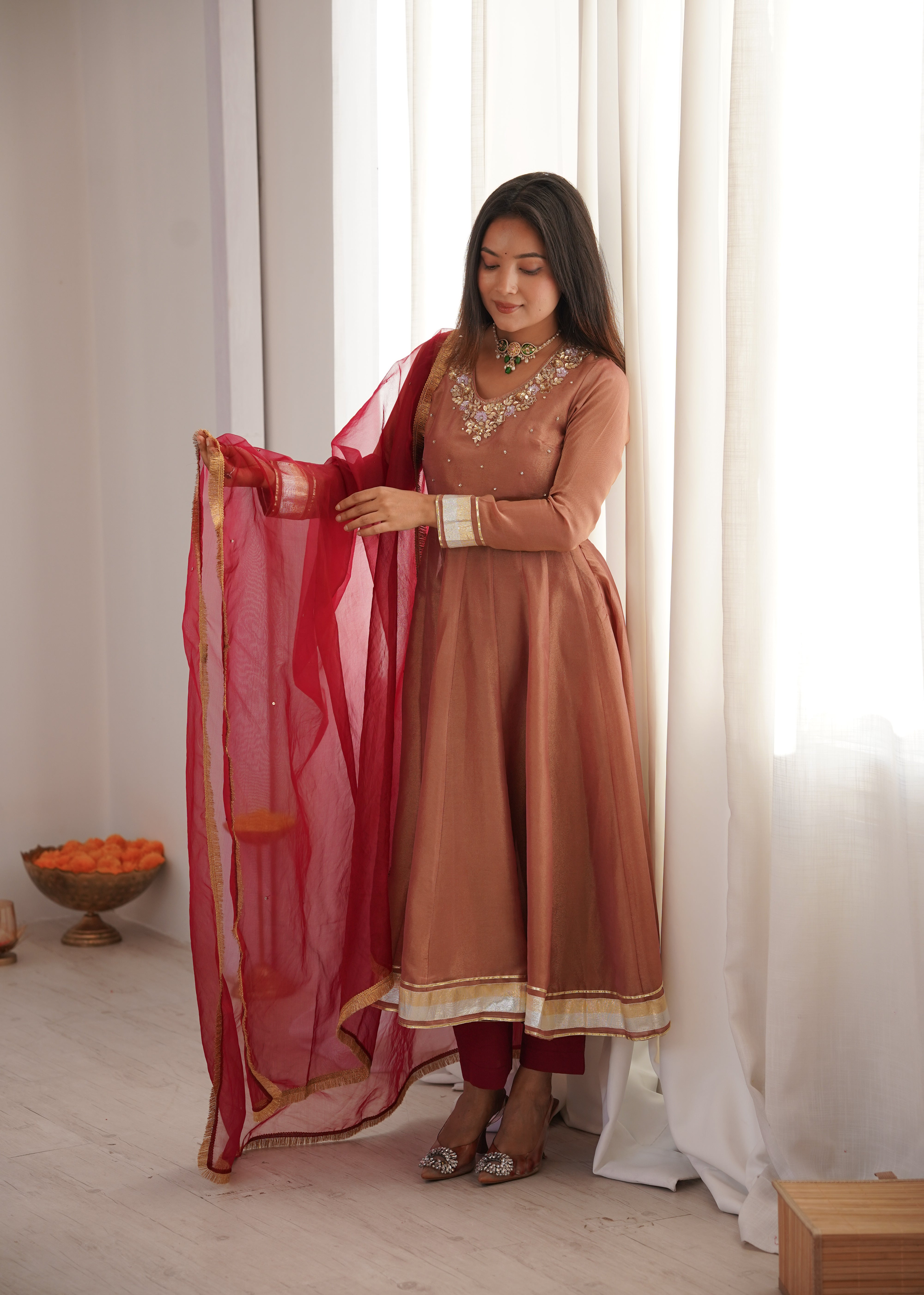 Woman in a traditional outfit holding a red dupatta against a white curtain background